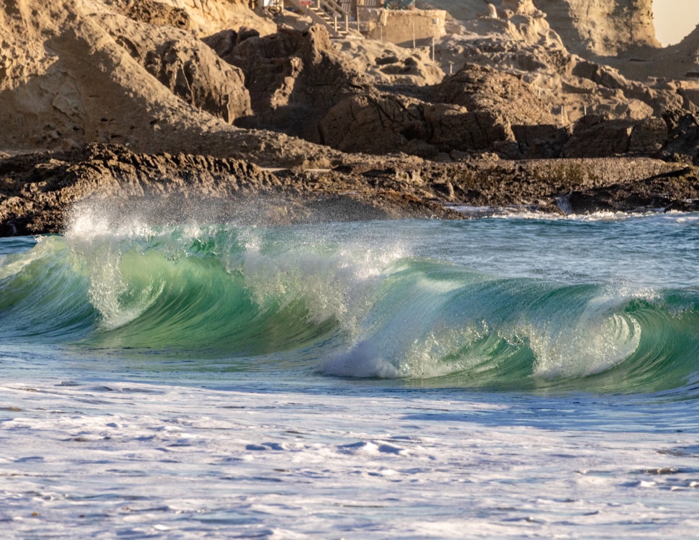 A green-blue wave breaking along a rocky Orange County coastline